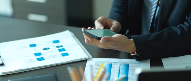 Крупным планом вид руки бизнесмена, использующей смартфон во время работы с деловой информацией на офисном столе