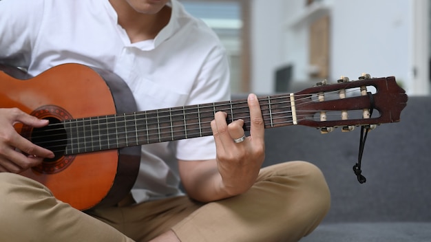 Close up view of casual man playing guitar while sitting on sofa at home.