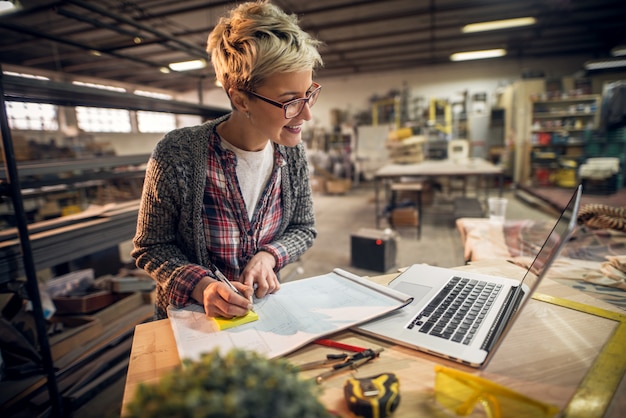 Close up view of charming smiling motivated short hair female engineer with eyeglasses working with blueprints and laptop in the workshop