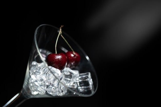 Close up view of cherry in a martini glass among ice in black background with flare.