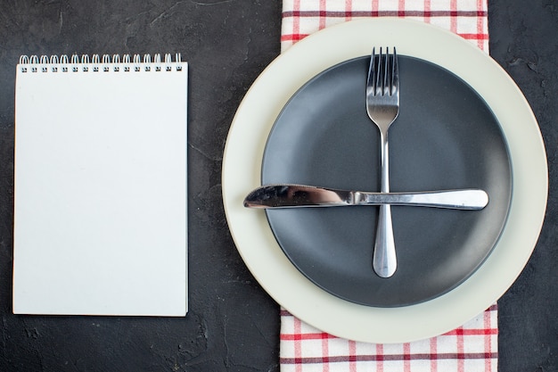 Close up view of cutlery set on dark gray color and white empty\
plates on red stripped towel next to notebook on black\
background