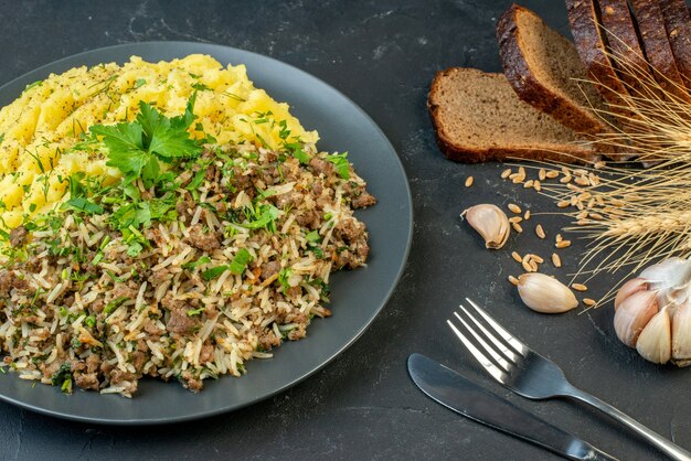 Крупным планом вид вкусного ужина с мясом и картофельным пюре на серой тарелке, ломтиками хлеба, набором столовых приборов с чесночным шипом на черном фоне