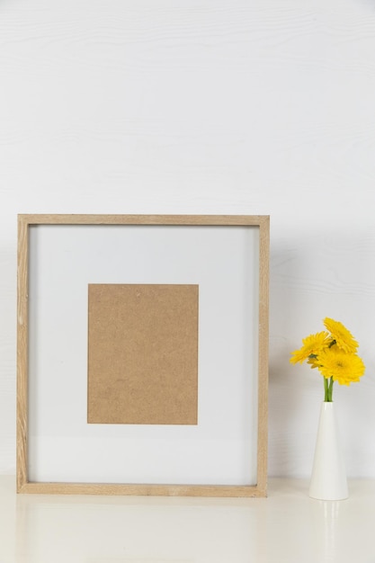 Photo close up view of an empty picture frame, with yellow flowers placed in a white vase arranged on a plain white background