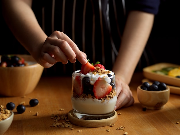 Vista ravvicinata della mano femminile decorazione fragola su una ciotola di muesli con yogurt greco e frutti di bosco
