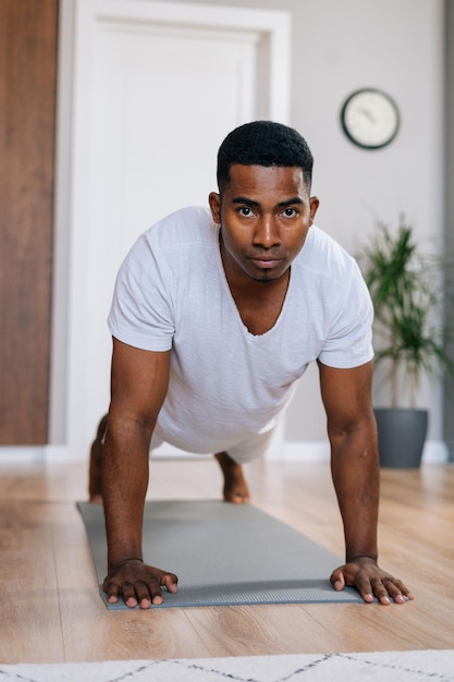 Close-up view of focused African-American man doing push-up on floor at bright domestic room, looking at camera . Concept of sport workout training at home gym.