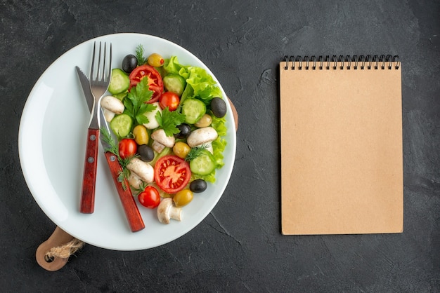 Close up view of fresh black and yellow olives chopped vegetables and cutlery set on a white plate notebook on black background with free space