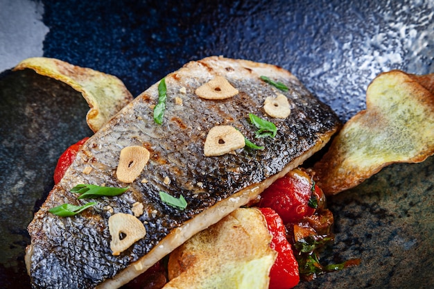 Close up view on fresh pan-fried seabass served with potato and tomato cherry in dark plate