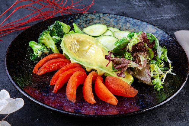 Close up view on green salad with avocado in served in black textured plate on dark. Flat lay food. Vegan fresh salad.