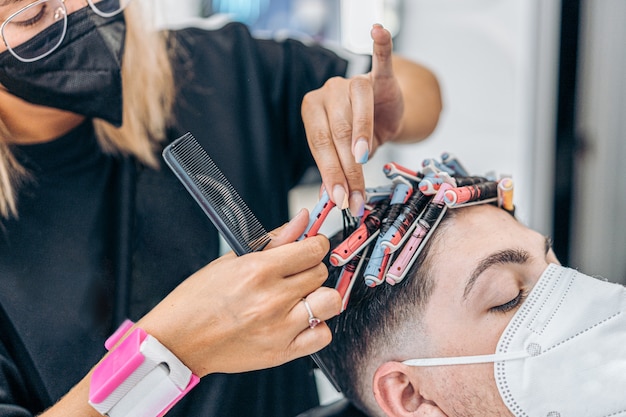 Close up view of a hairdresser curling the hair of a man with rollers while using mask