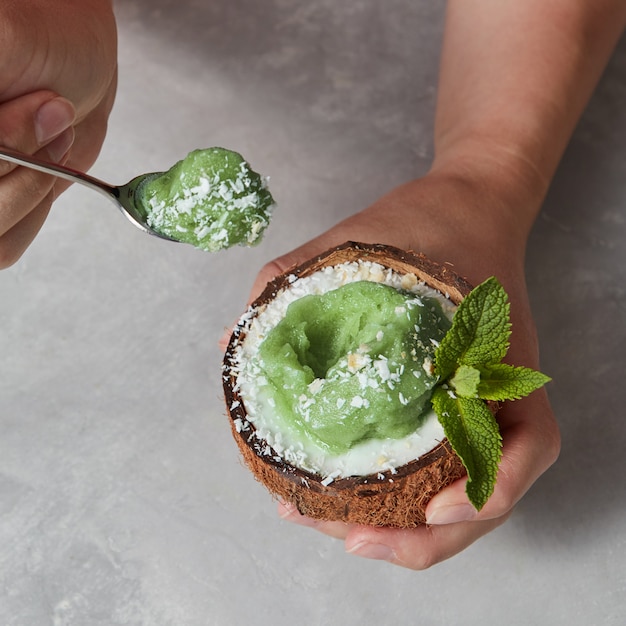 Vista ravvicinata una metà di cocco con gelato alla menta verde con un rametto di menta e scaglie di cocco nelle mani femminili sopra un tavolo grigio marmo.