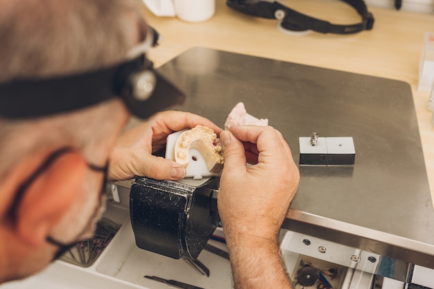 Vista ravvicinata delle mani di un maschio adulto caucasico che lavora su uno dei pezzi di uno stampo in ceramica in un laboratorio odontotecnico specializzato in ceramica ossea dentale per creare corone di porcellana