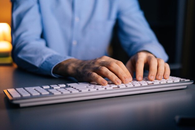 Close-up view of hands of unrecognizable man working typing on wireless keyboard computer. Male freelancer or student is working sitting on desk at home office at late night in dark room.