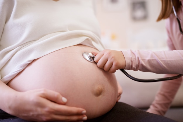 Photo close up view of little toddler girl checking health condition on the shirtless pregnant tummy of her mother with a stethoscope while resting at home.