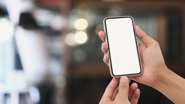 Close up view of man hands holding mock up smart phone with modern cafe blurred background.