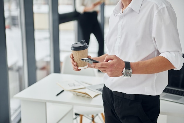 Photo close up view of man's hands with smartphone and cup of drink group of people is indoors in the office