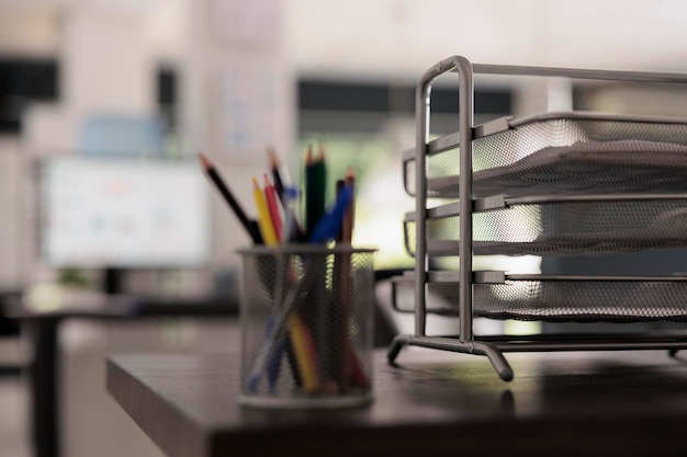 Close up view of metal pencil holder with colored pencils in\
creative design office, next to folder tray with project documents.\
out of focus shot of monitor on in background in empty\
workplace.