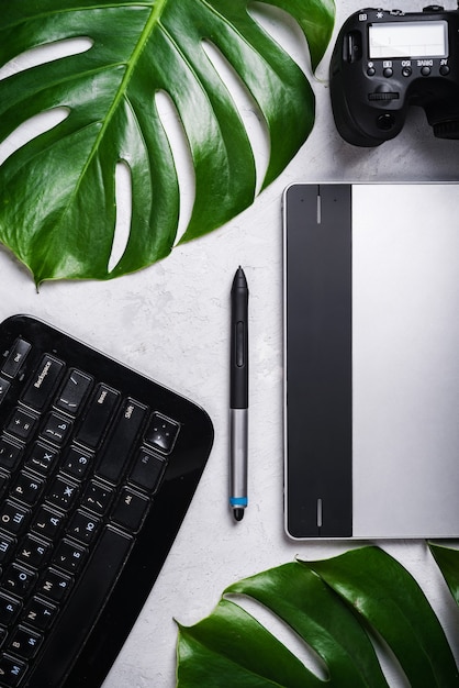 Close-up view of photographer\'s of graphic designer\'s\
workplace. tablet, stylus, camera, keyboard with russian and\
english letters, monstera green leaf.