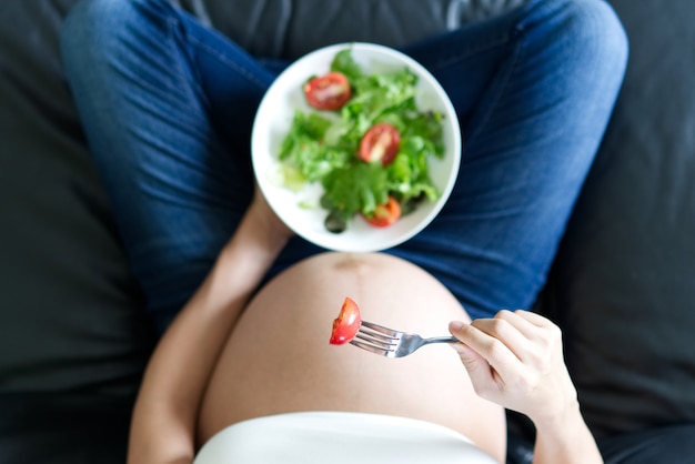 Close up view of pregnant women sitting on sofa holding salad bowl and eating tomato.