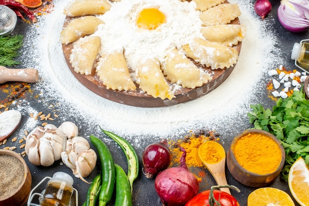 Close up view of raw dumplings on wooden tray around flour and set of foods on black surface