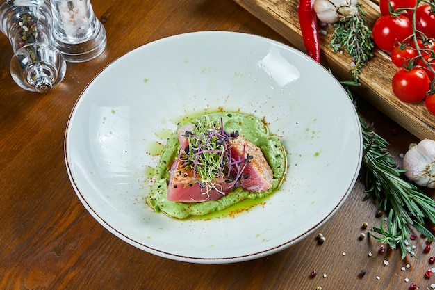 Close up view on roasted Medium-rare fried tuna with mashed avocado. Restaurant serving hot seafood in a white plate on a wooden table.