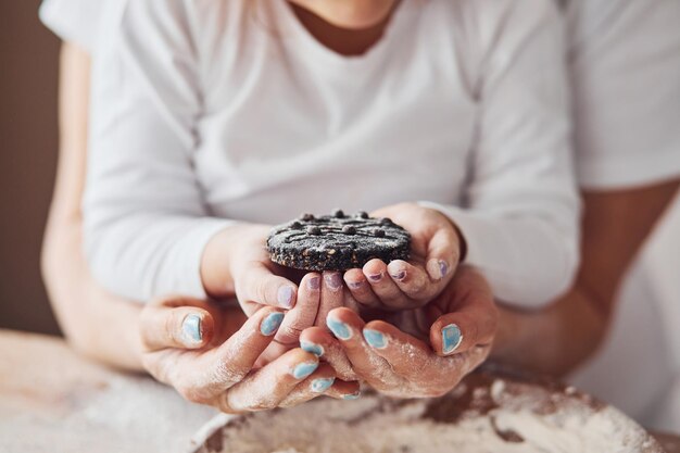 Close up view of senior woman with her granddaughter that holding fresh sweet cookie in hands on the kitchen with flour in plate on table