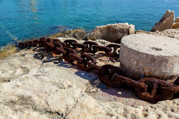 Close-up view of stone bollard with an old rusty chain on the blue water background.