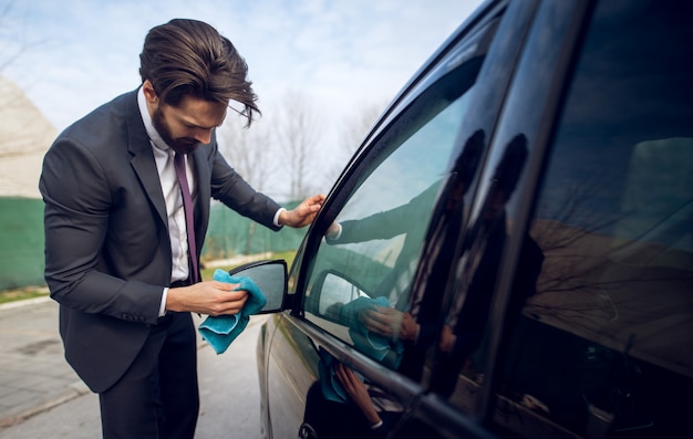 Close up view of stylish focused hardworking young businessman cleaning rearview mirror of his black car with a blue microfiber cloth.