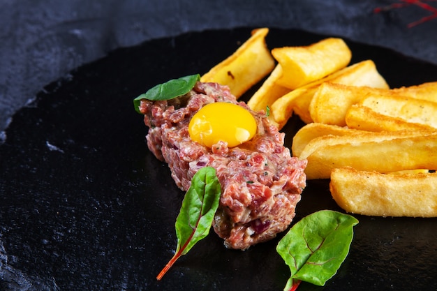 Close up view on tasty beef tartare steak served with egg yolk, potato dip on black stone plate on dark background.