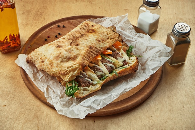 Photo close up view on tasty traditional italian sandwich panuozzo - oven-baked tortilla with tomatoes, melted mozzarella, minced beef and arugula on a wooden board. street food