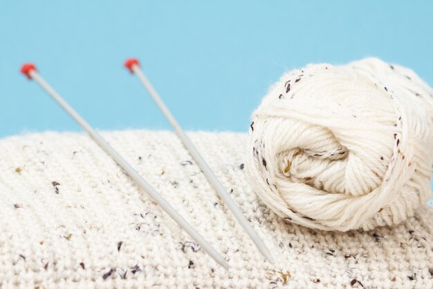 Close-up view of white skein of yarn and pullover with metal knitting needles on blue background. Knitting concept. Shallow depth of field.