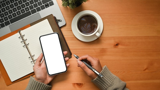 Close up view of woman holding pen and using smart phone on wooden desk Empty screen for your advertise design