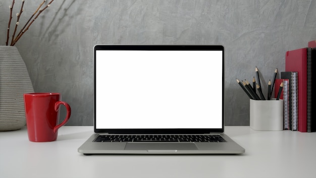 Foto chiuda sulla vista dell'area di lavoro con il computer portatile dello schermo in bianco, gli articoli per ufficio e la tazza di caffè rossa sulla tavola bianca con la parete grigia del sottotetto