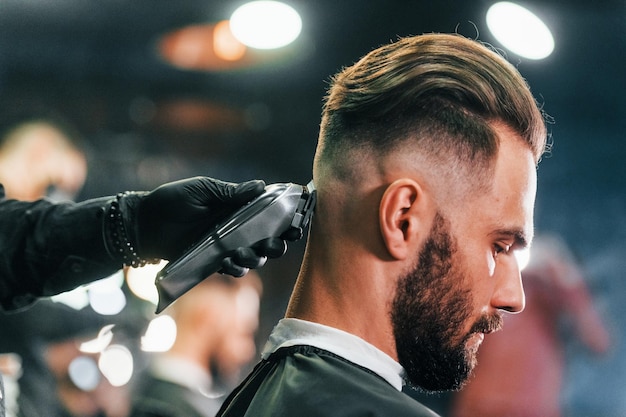 Close up view of young bearded man that sitting and getting haircut in barber shop