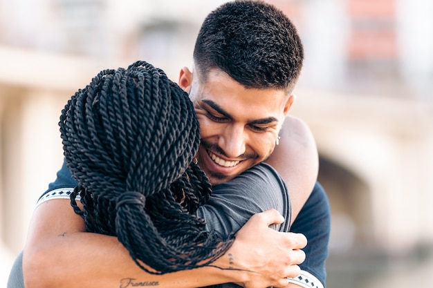 Close-up view of a young caucasian man hugging a young african woman while smiling in the street