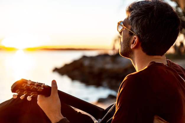 Close up View of Young Man Playing Guitar Outside whit Seaside as Background at Sunset Time