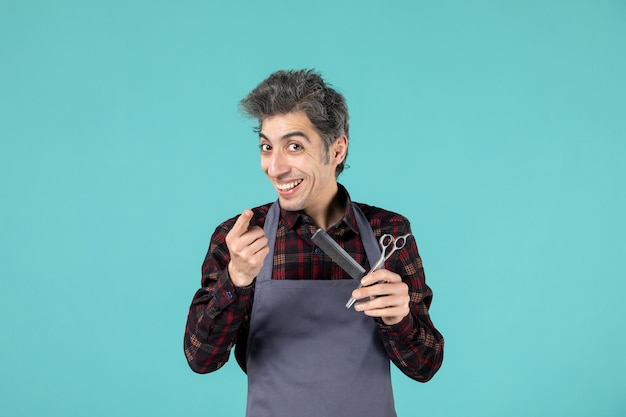 Крупным планом вид молодого улыбающегося мужского парикмахера в сером фартуке и держащего ножничную расческу, направленную вперед на пастельно-синем цветном фоне