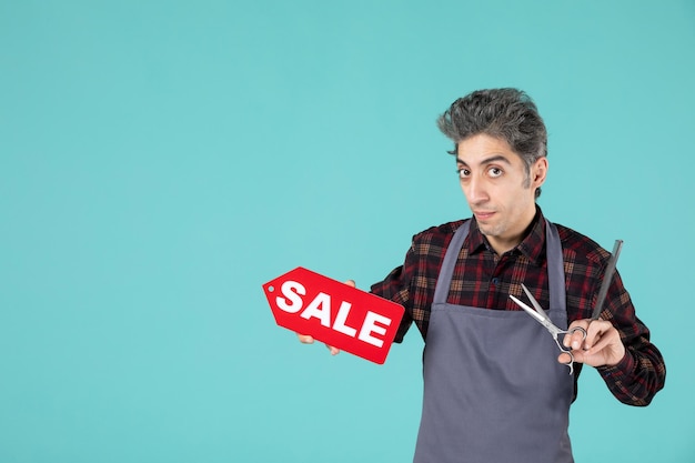 Close up view of young unsatisfied male barber wearing gray apron and holding scissors and comb showing sale icon on soft blue color surface