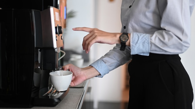 オフィスの休憩時間中にコーヒーマシンでコーヒーを作る若い女性のクローズアップビュー。