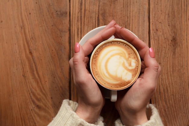 Close-up vrouwelijke handen met kopje koffie cappuccino met schuim met mooi patroon. Perfecte rode gellak manicure. Houten natuurlijke vintage tafel. Creatieve kleur warme nabewerking