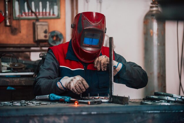 防護服を着た溶接工をクローズアップし、工業用ワークショップの後ろにいる他の労働者と一緒に工業用テーブルで金属パイプを溶接するマスク