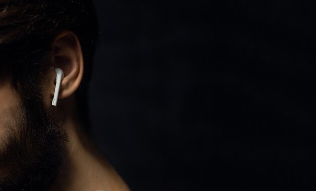 Close-up of wireless earphone in male ear on wall of black color with copy space.