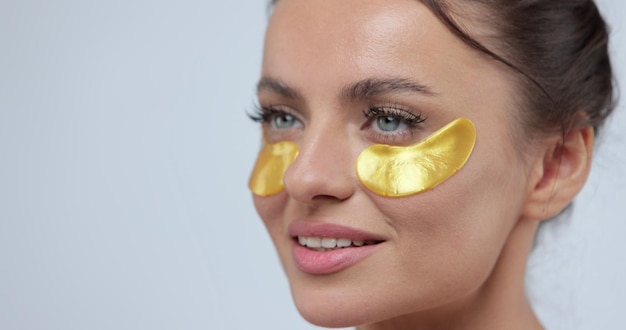 Close up of woman applying gold eye patches sensually posing on camera over gary background Beauty concept