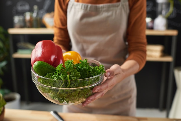 彼女がサラダを調理しようとしている新鮮な野菜とボウルを保持しているエプロンの女性のクローズアップ