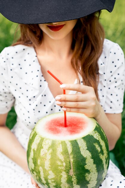 Photo close up of woman in black hat holds a whole watermelon with cocktail straw.