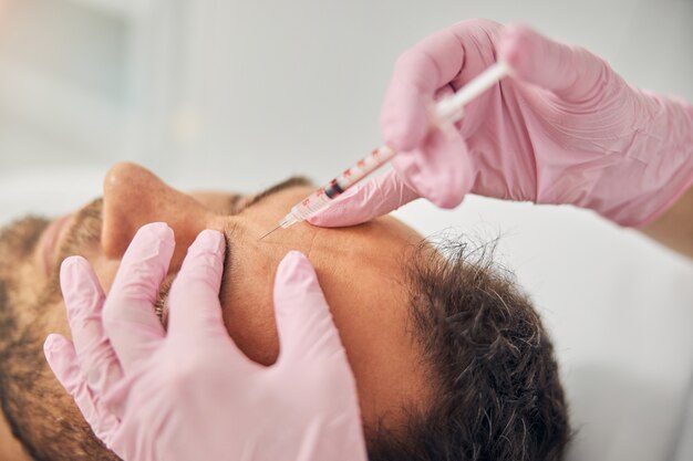 close up of woman cosmetologist in sterile gloves making injection in young man forehead