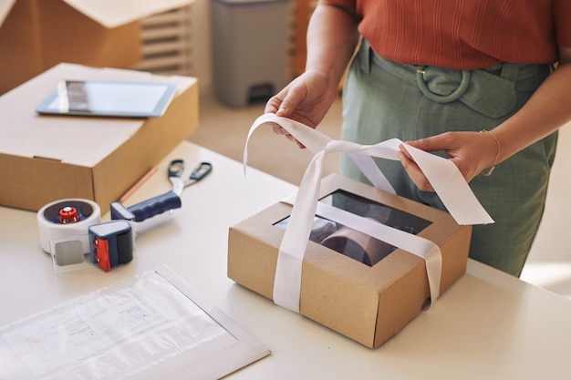 Close-up of woman decorating cardboard box with ribbon at the table before delivery