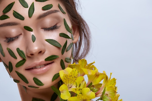 Close-up of woman face full of green leaves with yellow flowers isolated on grey