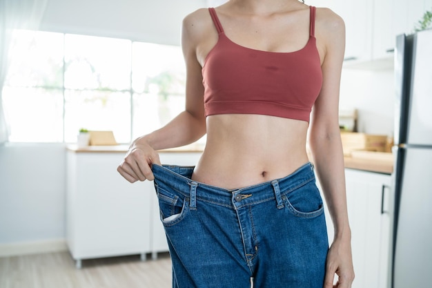 Close up of woman feel happy after lose weight for health in house Attractive beautiful skinny girl wearing old big jeans with abs abdomen sick packs in kitchen at home Healthy food and Diet concept