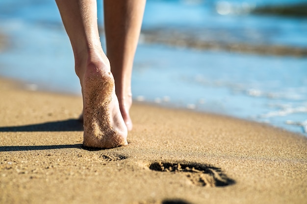 Chiuda in su dei piedi della donna che cammina a piedi nudi sulla sabbia lasciando impronte sulla spiaggia dorata. vacanze, viaggi e concetto di libertà. persone che si rilassano in estate.