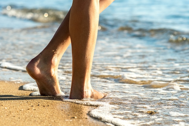 Close up of woman feet walking barefoot on sand leaving footprints on golden beach.